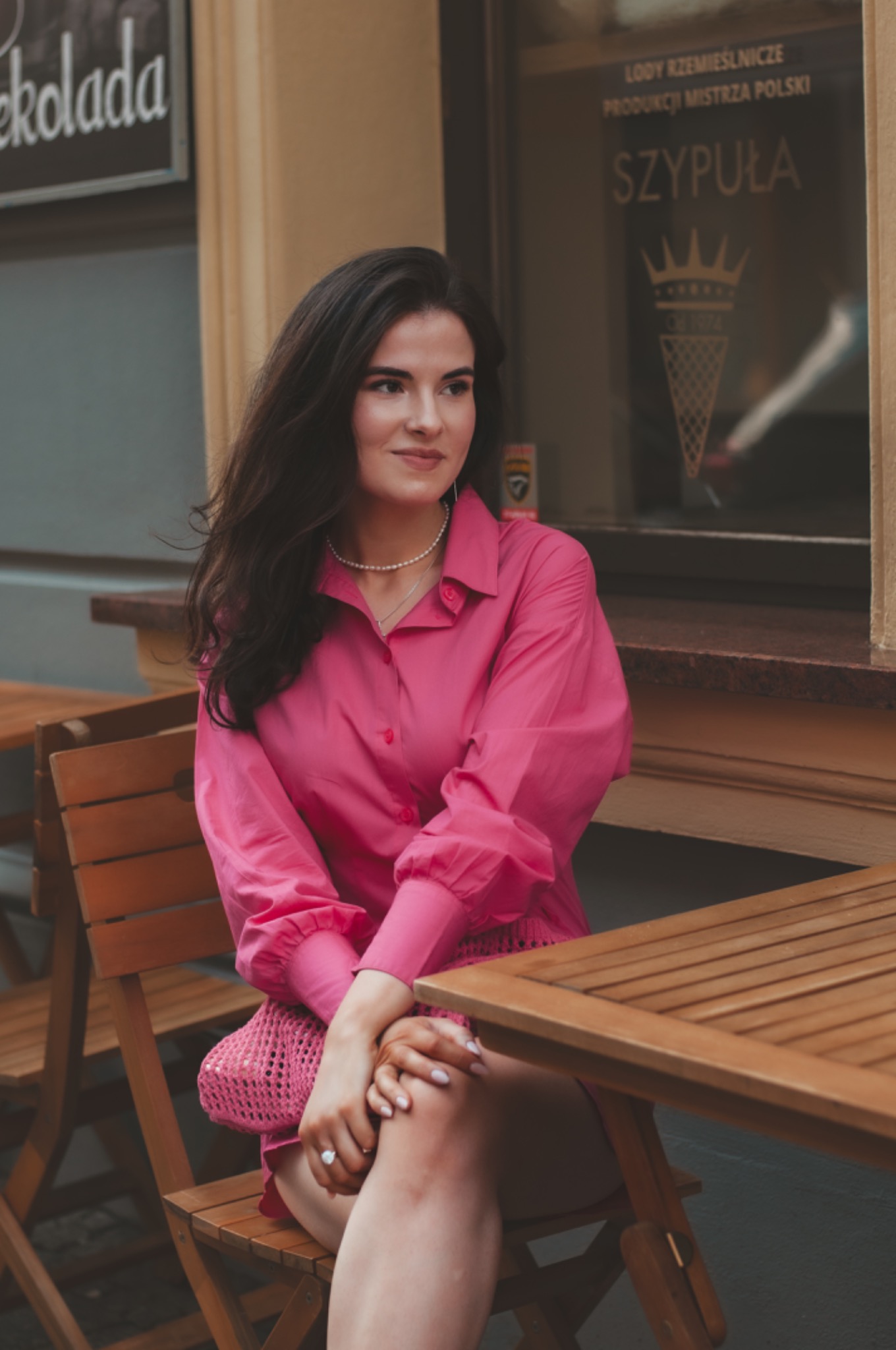 Portret kobiety w różowej koszuli siedzącej przy drewnianym stoliku kawiarnianym na zewnątrz, z widocznym szyldem lodziarni w tle.