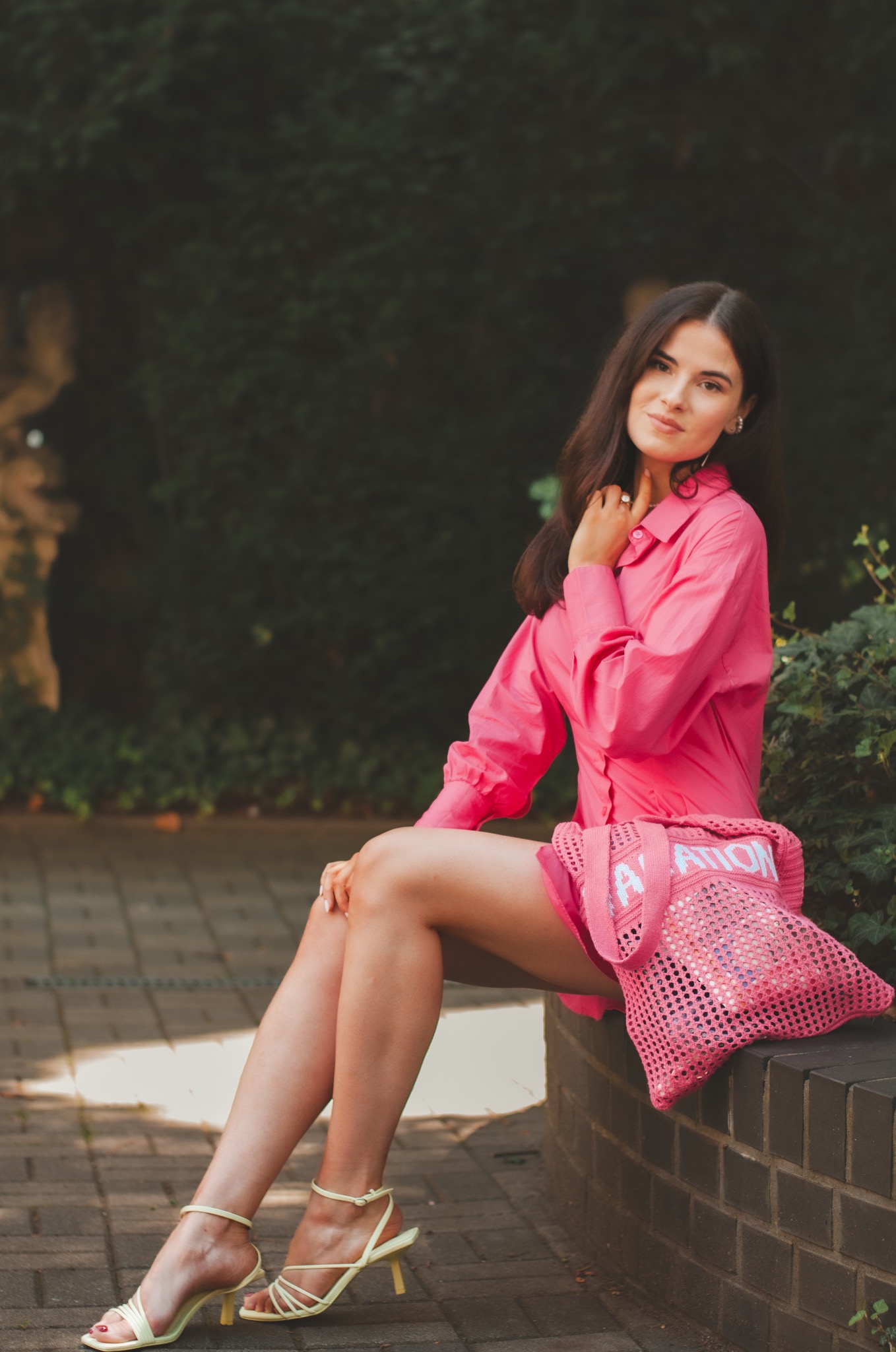 Portret kobiety w różowej koszuli i z torebką na tle zieleni w Murów, siedzącej na murku.