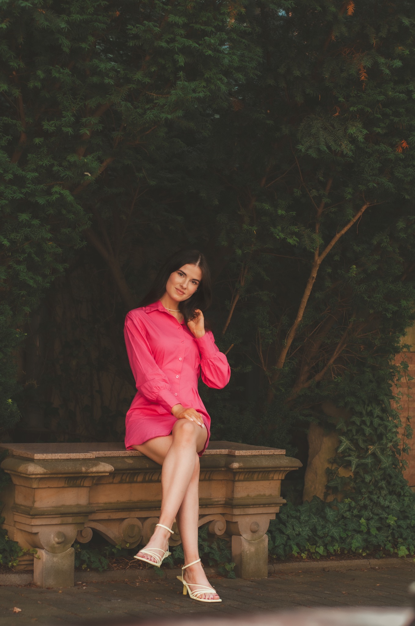 Portret kobiety w różowej sukience koszulowej i jasnych sandałach siedzącej na kamiennej ławce w ogrodzie.