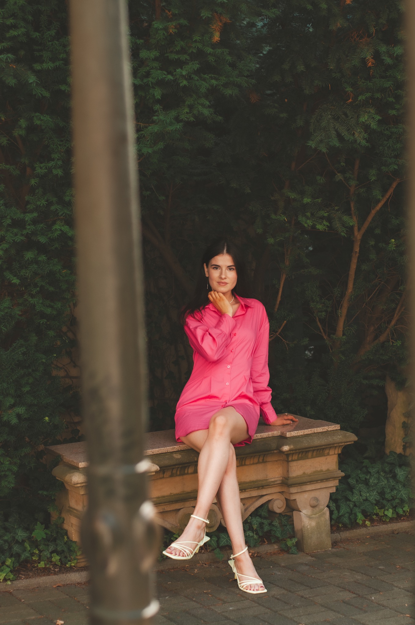 Kobieta w różowej koszuli siedzi na kamiennej ławce w parku, pozując z ręką pod brodą, widok przez metalowe pręty.