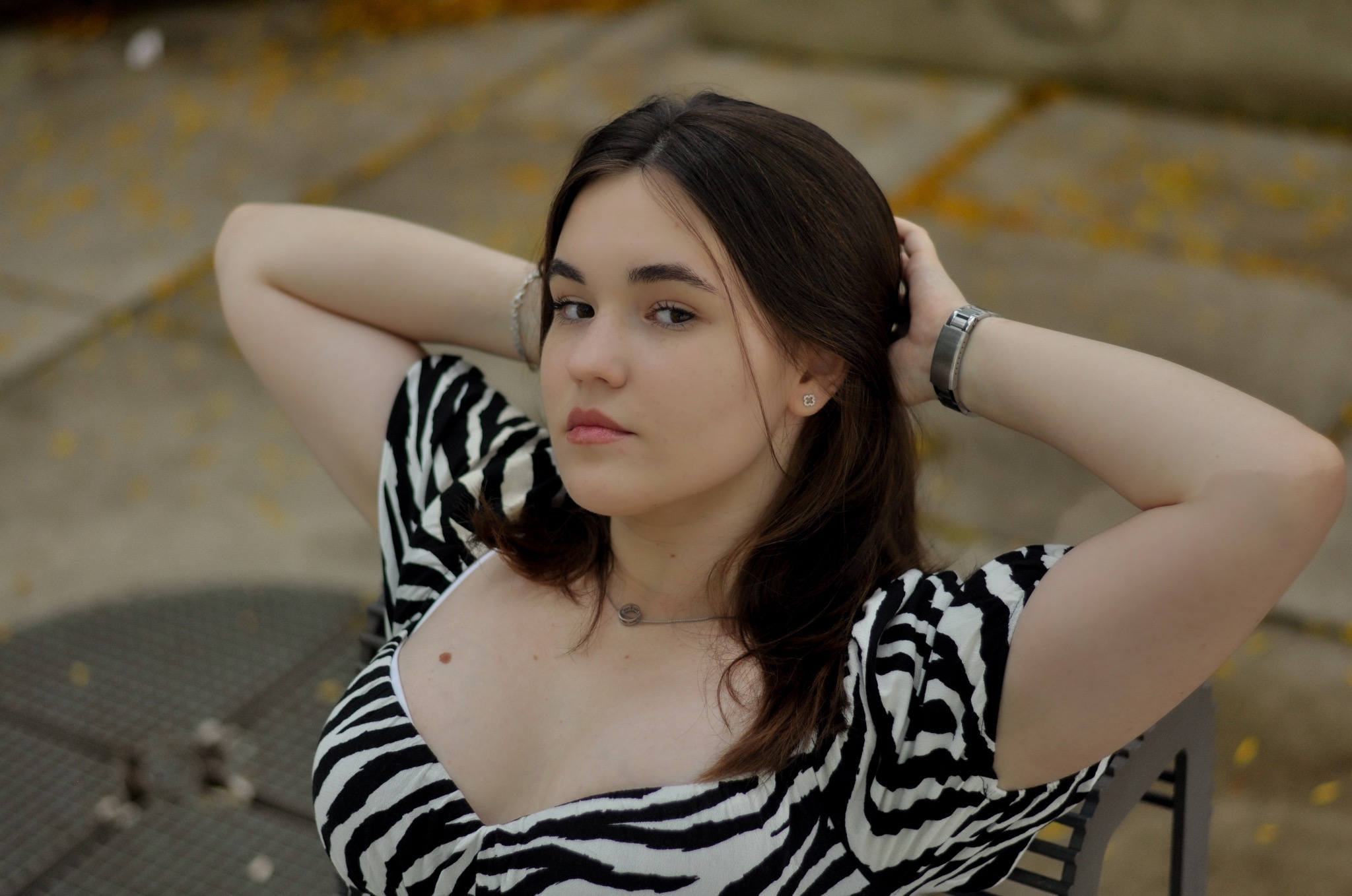 Portret młodej kobiety w bluzce w zebrę, pozującej z rękami uniesionymi za głową, zegarkiem na nadgarstku i delikatną biżuterią na szyi i uszach, na tle betonowej nawierzchni.