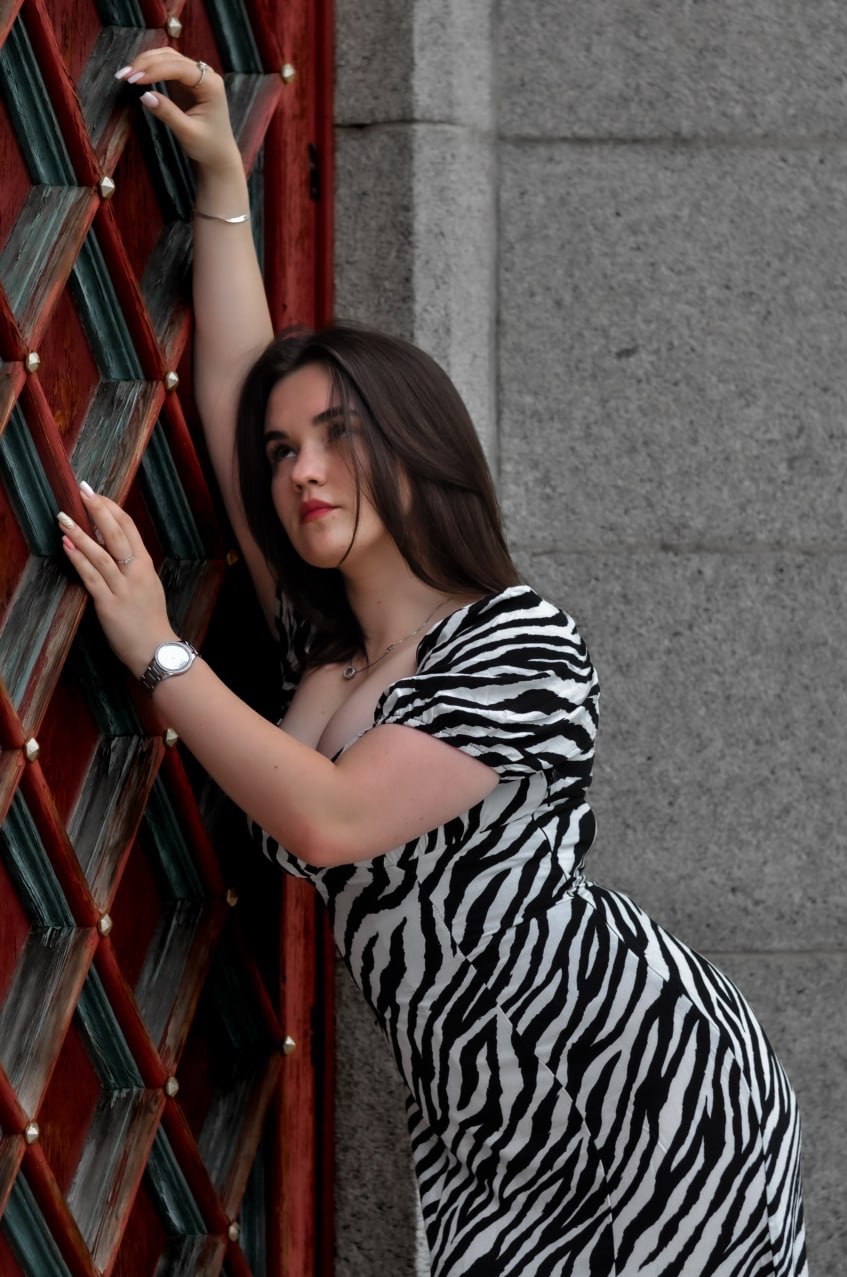 Portret kobiety w sukience w zebrę, opierającej się o ozdobne, drewniane drzwi z metalowymi elementami, na tle szarej, kamiennej ściany.