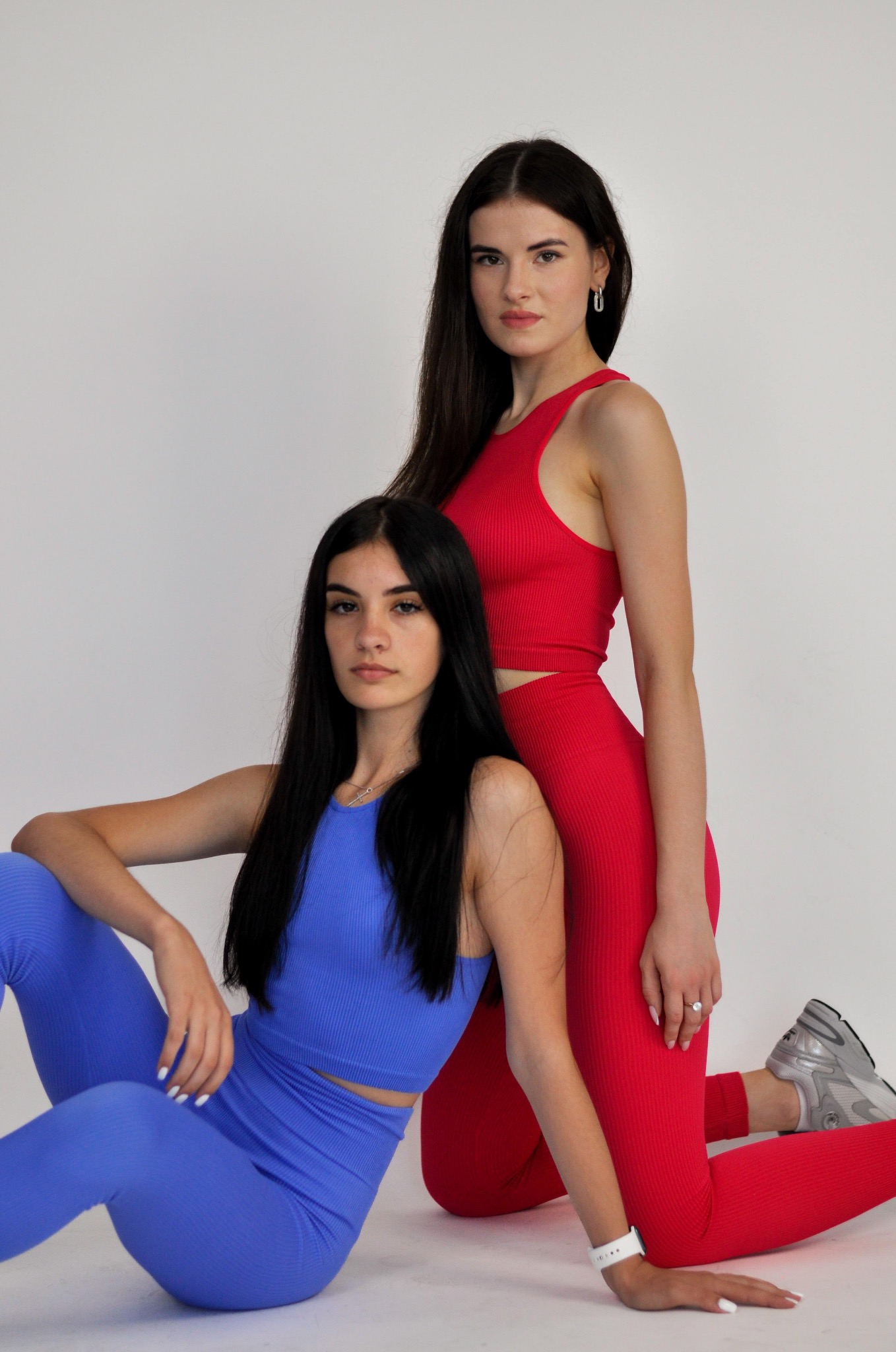 Dwie kobiety w sportowych kompletach: siedząca w niebieskim prążkowanym zestawie i stojąca w czerwonym prążkowanym zestawie, na białym tle.