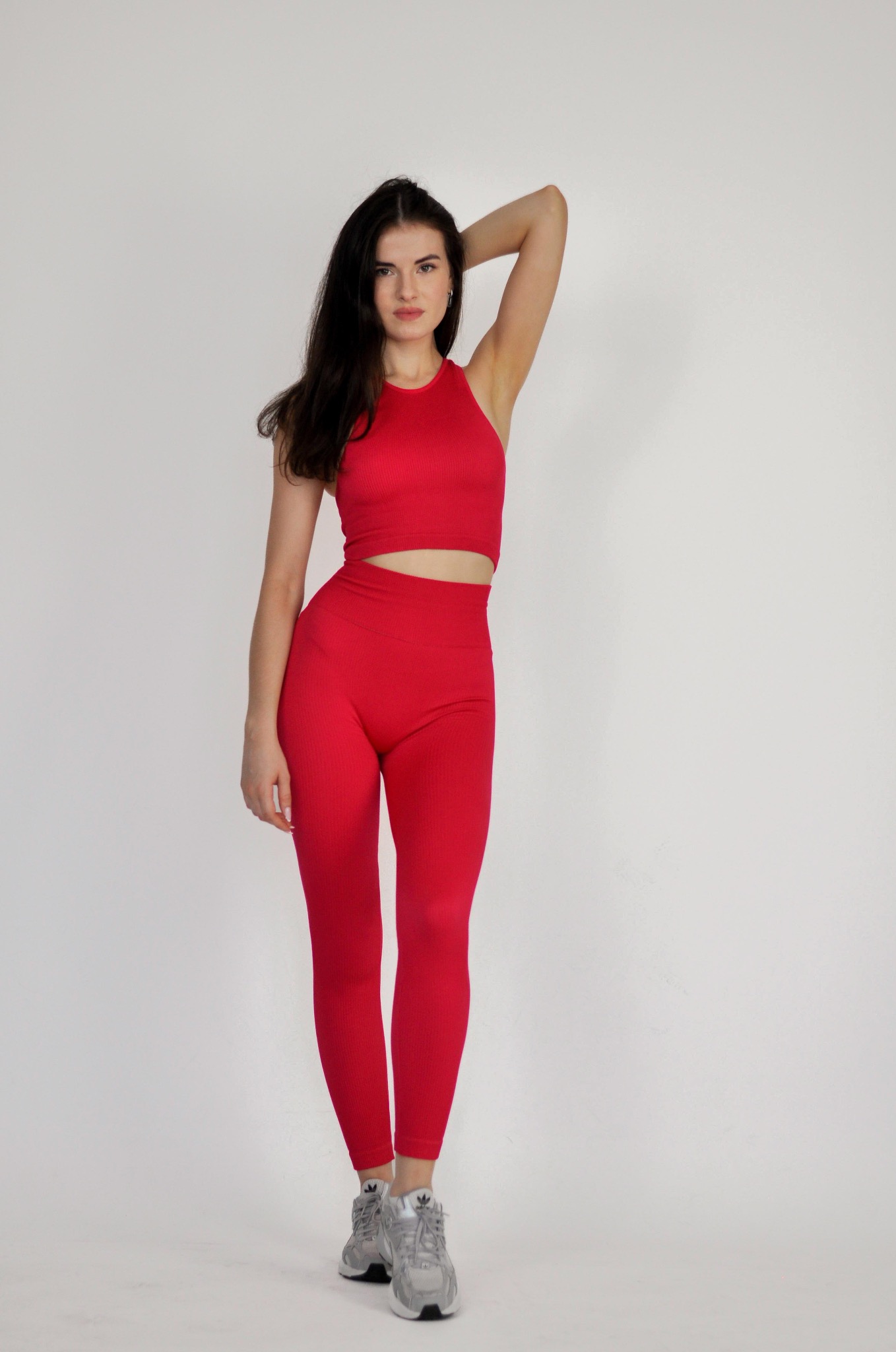 Portret kobiety w czerwonym sportowym stroju, pozującej na jasnym tle, z ręką uniesioną za głową i srebrnymi butami sportowymi.