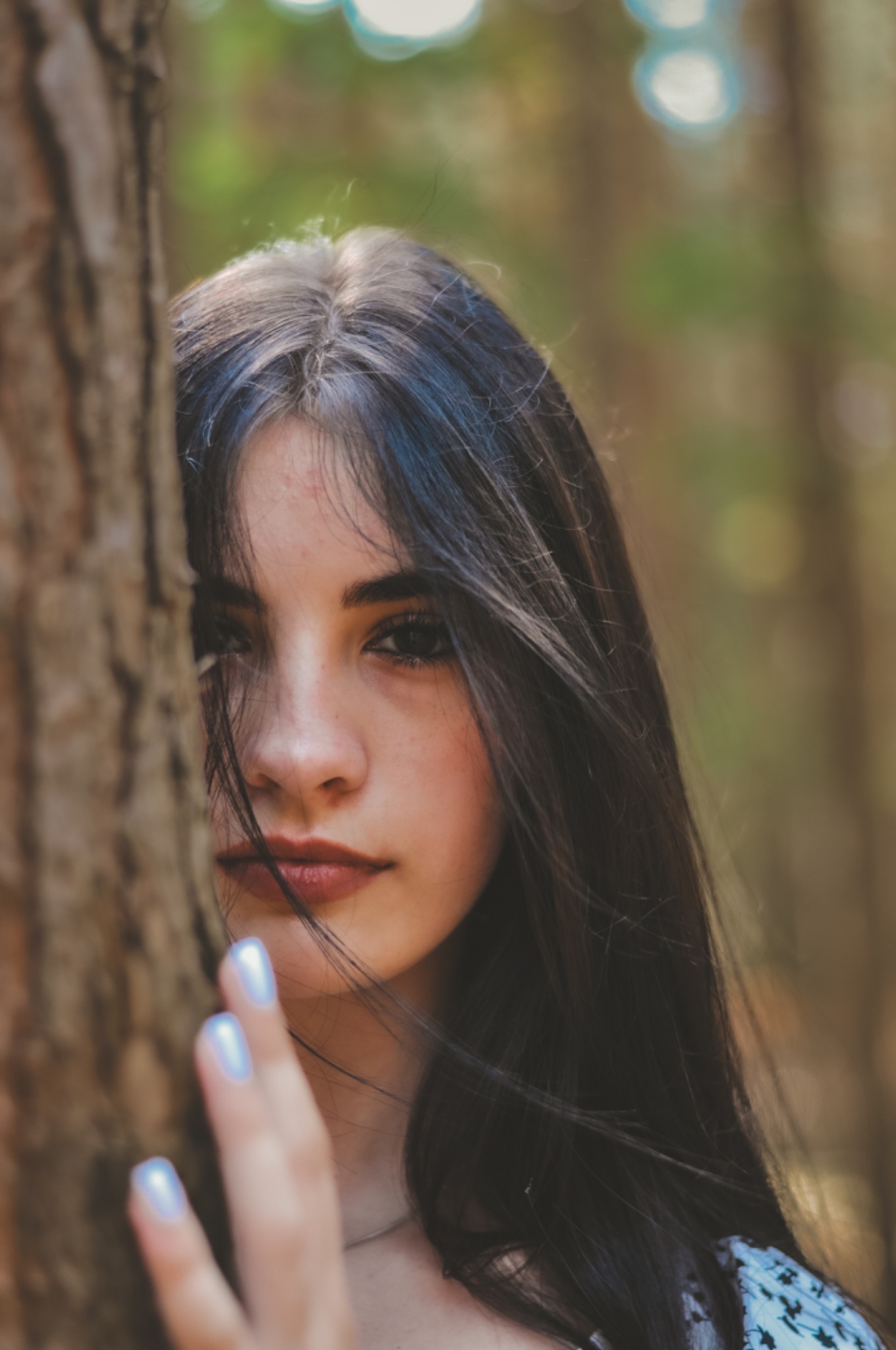 Portret młodej kobiety o ciemnych włosach, częściowo zasłoniętej przez drzewo, z delikatnym makijażem i opalizującym lakierem na paznokciach.