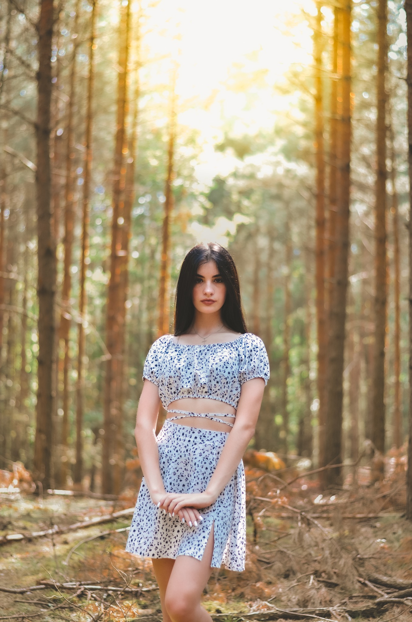 Portret młodej kobiety w kwiecistym komplecie, stojącej w lesie iglastym z promieniami słońca prześwitującymi przez korony drzew.