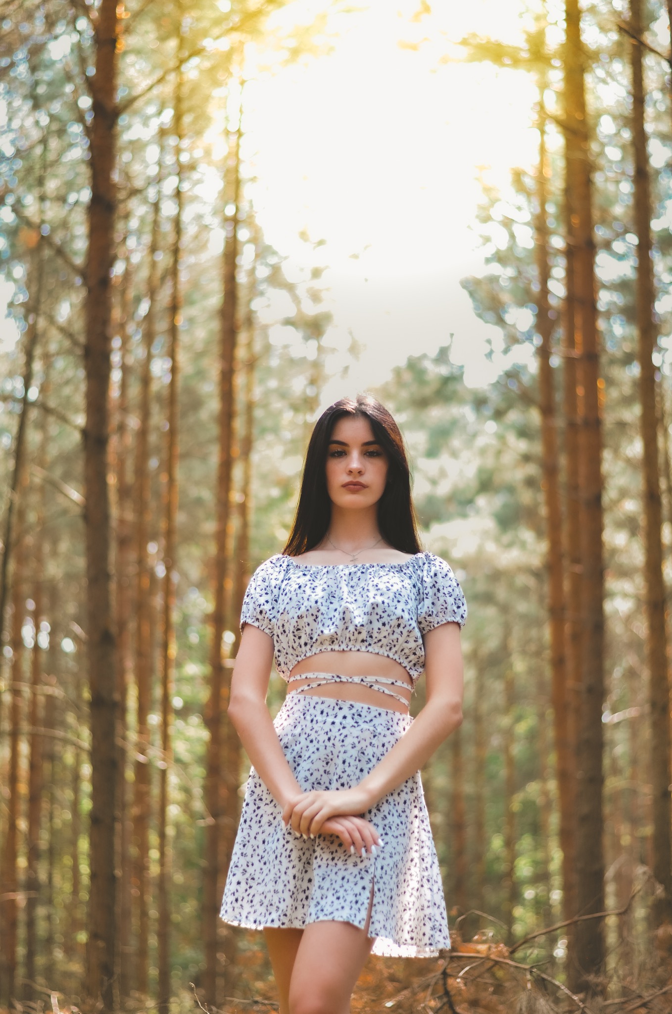 Portret młodej kobiety w kwiecistym stroju, stojącej na tle lasu z promieniami słońca przebijającymi przez korony drzew.
