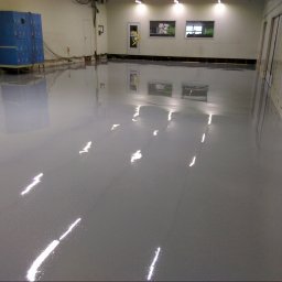 MARBUZ EPOXY ALEKSANDRA MATUSZEWSKA - Błyszcząca, szara posadzka w hali magazynowej, odbijająca światło lamp sufitowych. W tle widoczne niebieskie szafki i białe ściany.