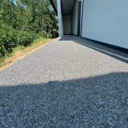 Szara posadzka żywiczna z kamyczków na zewnątrz budynku, wykonana przez firmę z Wińska. Widok z perspektywy chodnika, w tle zieleń i budynek.