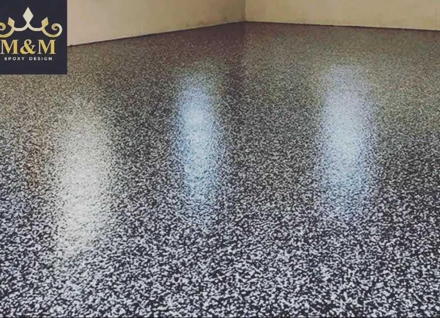 Błyszcząca, szaro-czarna podłoga z drobnymi plamkami, widoczny fragment ściany w rogu pomieszczenia, w lewym górnym rogu logo firmy M&M Epoxy Design.
