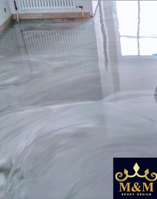 Błyszcząca, szara podłoga z efektem marmuru, widoczny grzejnik i okno w tle, w prawym dolnym rogu logo firmy M&M Epoxy Design.