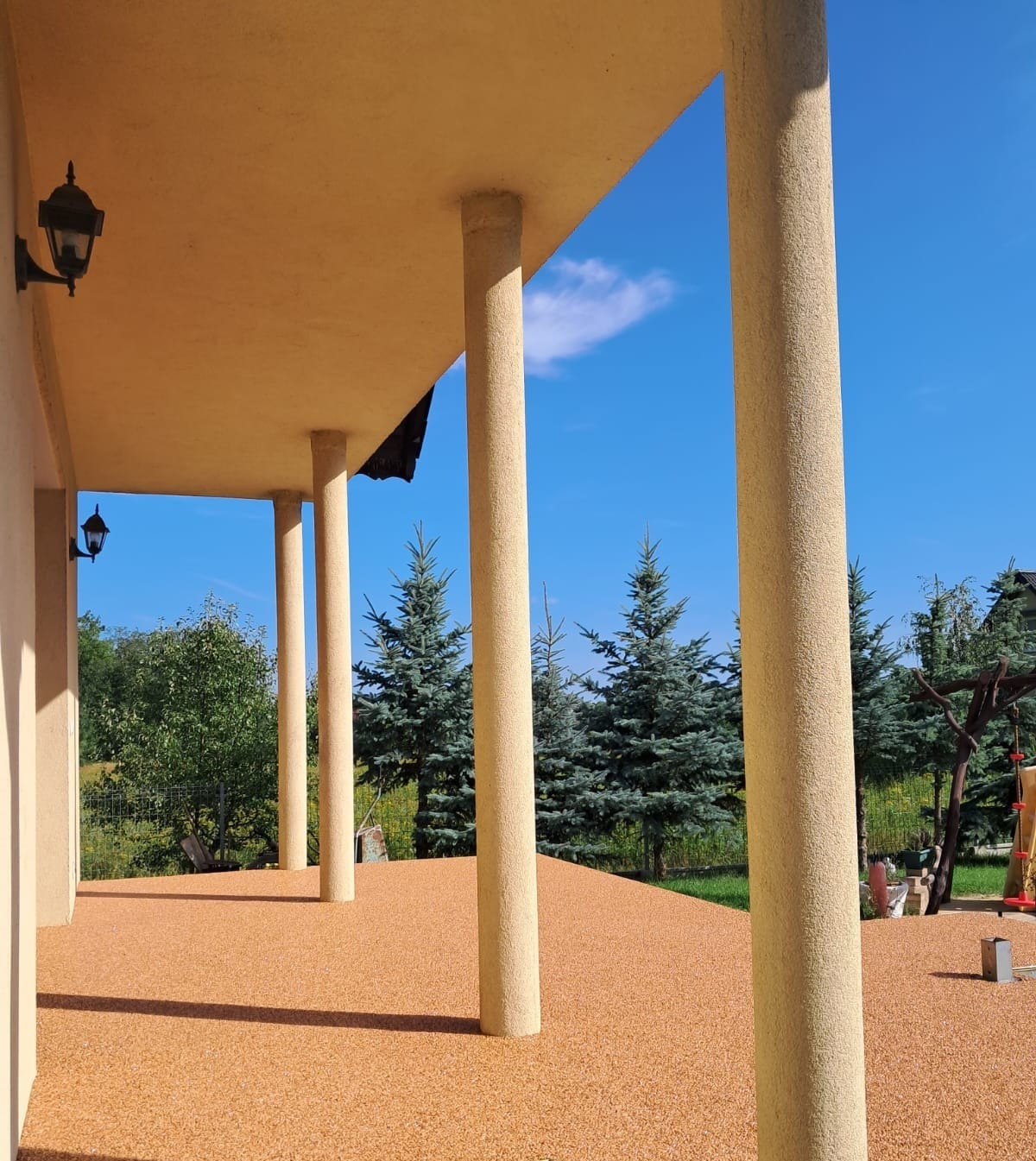 Zadaszony taras z beżowymi kolumnami i pomarańczową posadzką żywiczną na zewnątrz domu, w tle iglaki i niebieskie niebo.