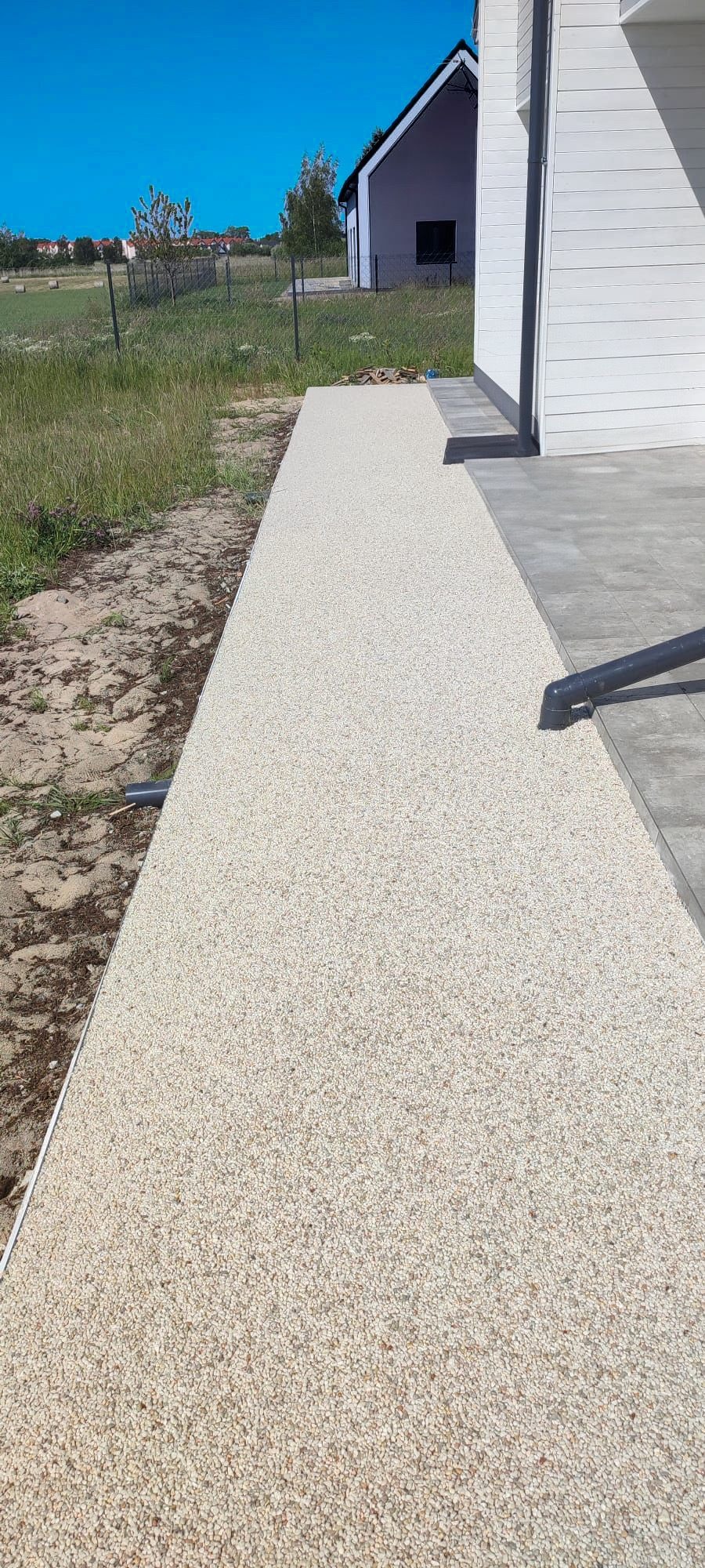 Beżowa powierzchnia z drobnych kamyczków, tworząca ścieżkę obok białego domu z szarą podmurówką, widok z góry w słoneczny dzień.