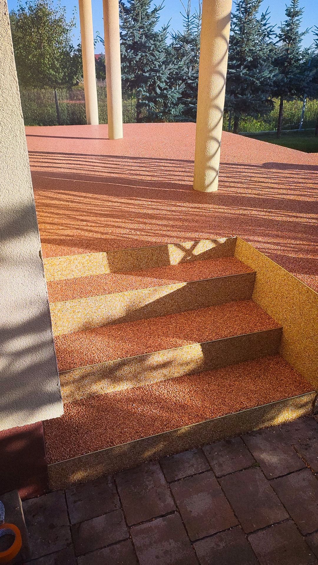 Schody i taras pokryte żywiczną posadzką w odcieniach pomarańczy i żółci, widoczne kolumny i iglaki w tle.