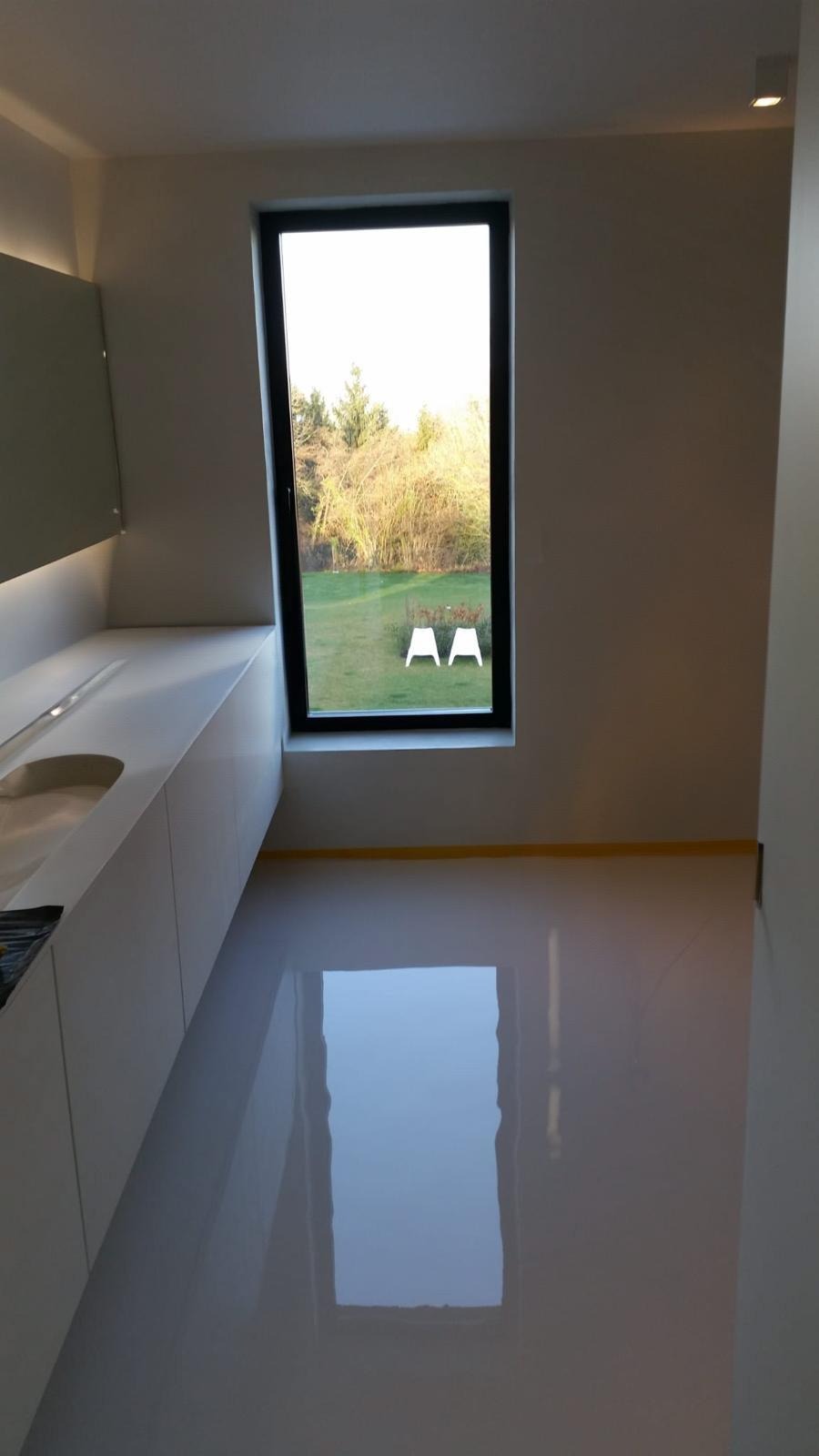 Błyszcząca, jasnoszara podłoga w nowoczesnym wnętrzu z dużym oknem, przez które widać trawnik i dwa białe krzesła. Żółta taśma zabezpieczająca na styku podłogi i ściany.