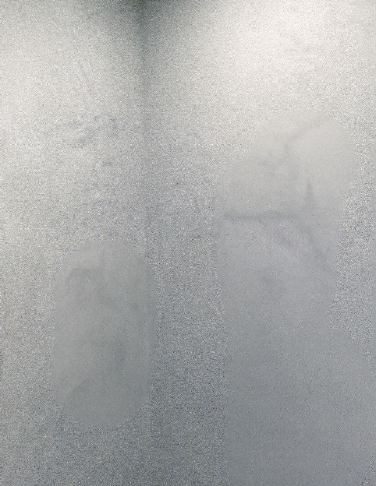 Ściana w rogu pomieszczenia pokryta szarą, dekoracyjną powłoką z subtelnymi, jaśniejszymi smugami i delikatnym połyskiem.