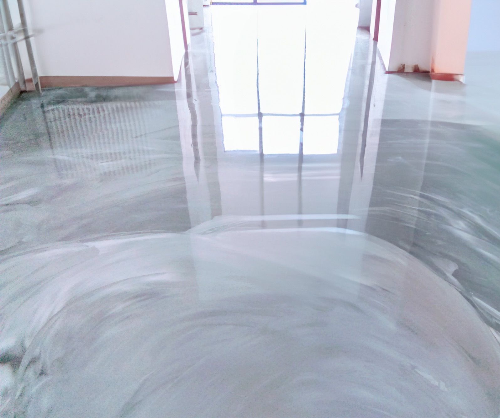 Błyszcząca, jasnoszara posadzka z efektem chmurkowania, widoczna w pustym pomieszczeniu z refleksami światła dziennego wpadającego przez okno.