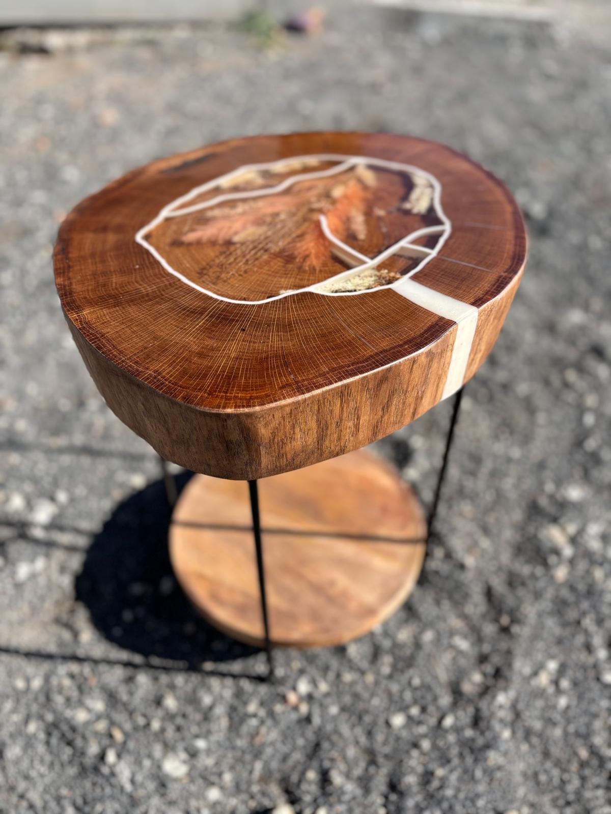 Stolik kawowy z blatem z przekroju pnia drzewa, wypełnionym żywicą epoksydową i fragmentami drewna, na metalowych nogach z dodatkowym drewnianym blatem na dole, sfotografowany na tle szarego żwiru.