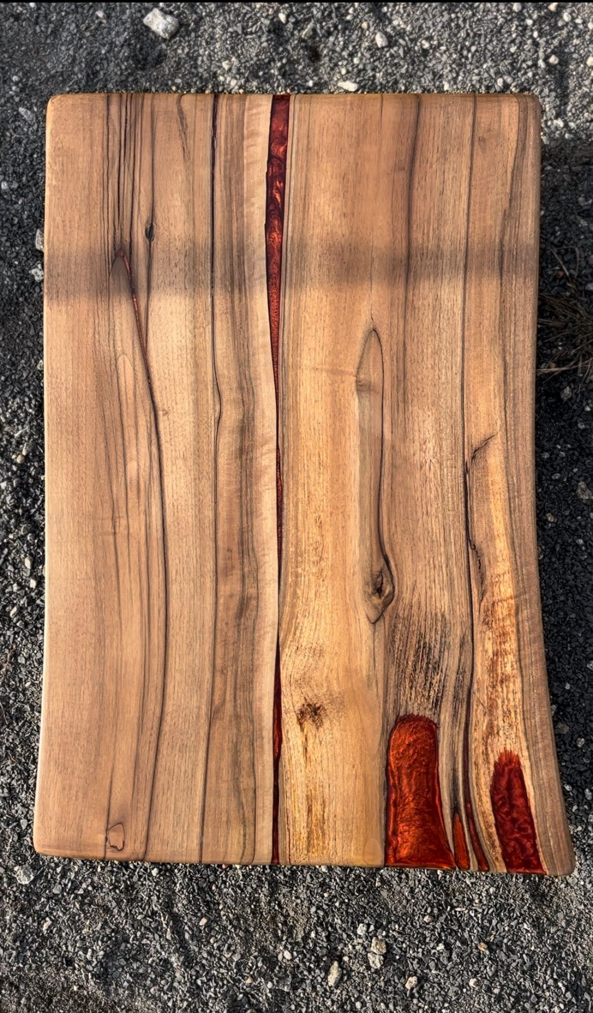Deska z drewna orzechowego z żywicą epoksydową w kolorze czerwonym, leżąca na szarym żwirze.