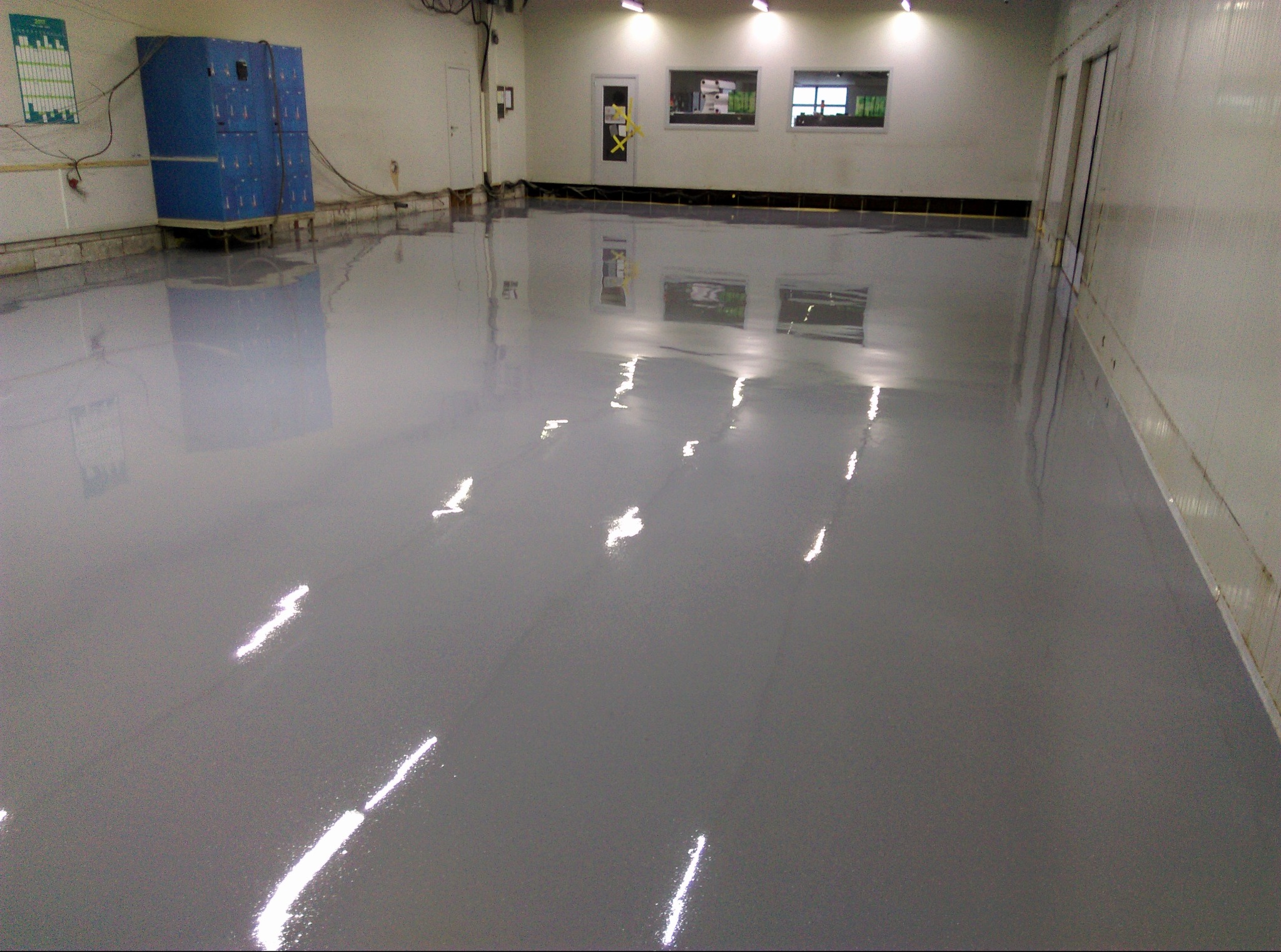 Błyszcząca, szara posadzka w hali magazynowej, odbijająca światło lamp sufitowych. W tle widoczne niebieskie szafki i białe ściany.