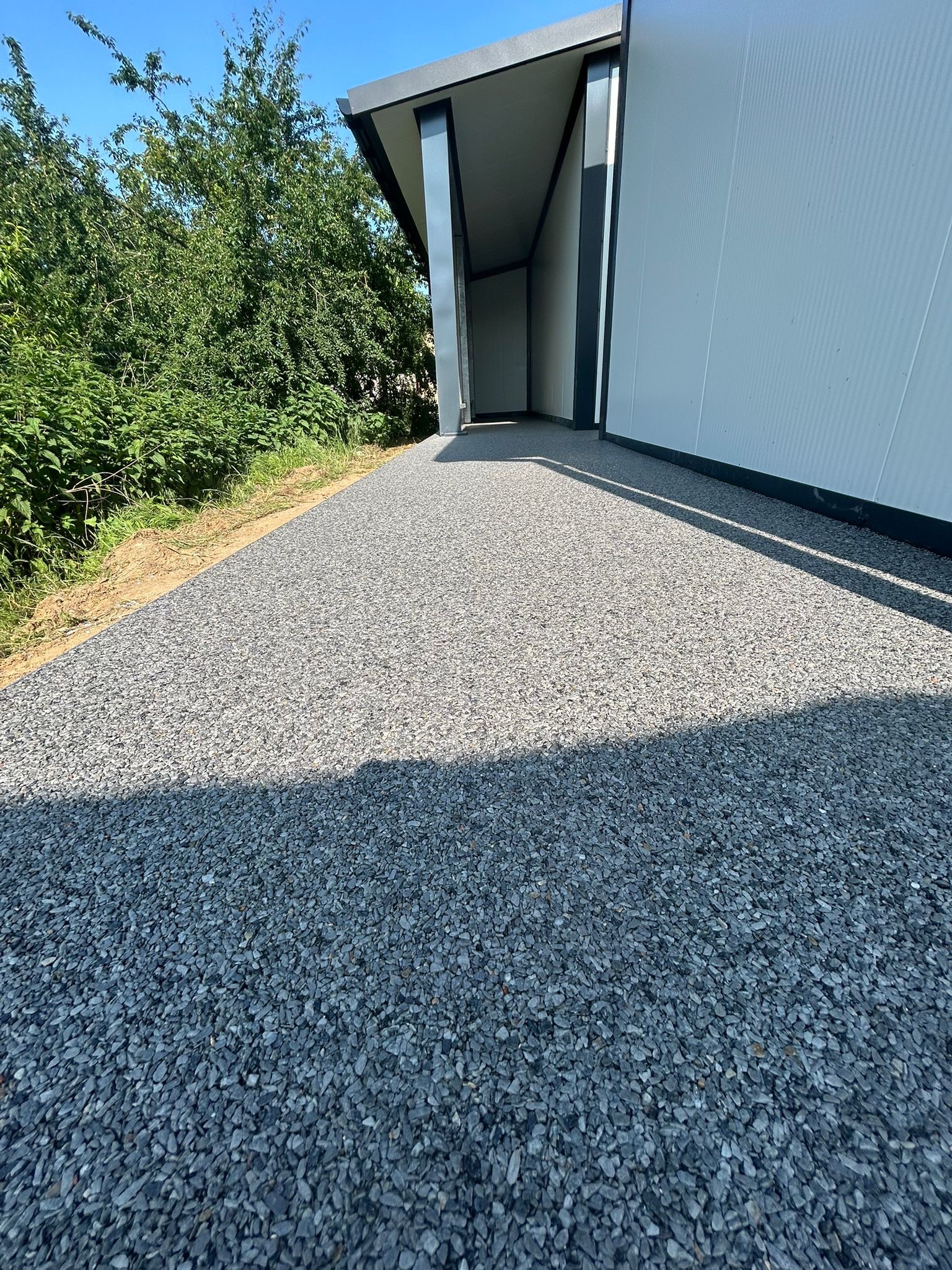 Szara posadzka żywiczna z kamyczków na zewnątrz budynku, wykonana przez firmę z Wińska. Widok z perspektywy chodnika, w tle zieleń i budynek.