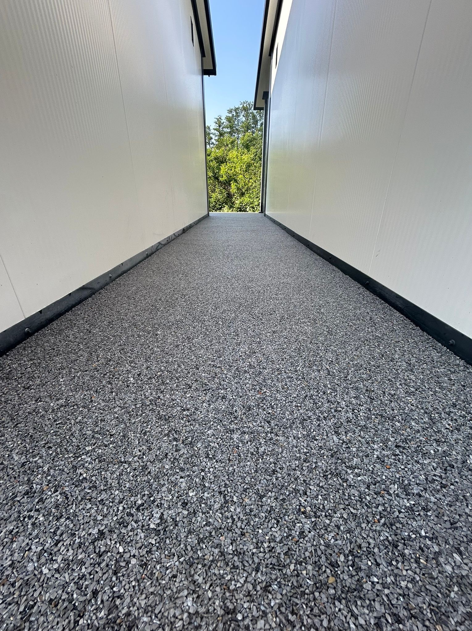 Szara, drobnoziarnista posadzka przemysłowa w wąskim korytarzu między białymi ścianami. Widok w perspektywie z zielenią w oddali.