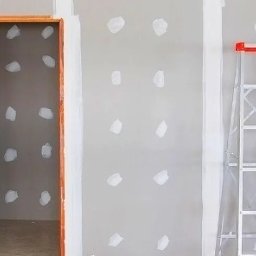 Renowacja Łukaszewicz - Ściana z płyt gipsowo-kartonowych po nałożeniu pierwszej warstwy szpachli w miejscach łączeń i po wkrętach, widoczna drabina aluminiowa z pomarańczowym elementem na górze i futryna drzwiowa...