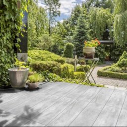 Renowacja Łukaszewicz - Szare płytki tarasowe ułożone na zewnątrz, z doniczkami z roślinami i widokiem na bujny ogród z drzewami i krzewami.
