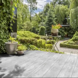 Szare płytki tarasowe ułożone na zewnątrz, z doniczkami z roślinami i widokiem na bujny ogród z drzewami i krzewami.