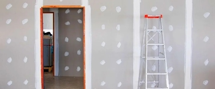 Ściana z płyt gipsowo-kartonowych po nałożeniu pierwszej warstwy szpachli w miejscach łączeń i po wkrętach, widoczna drabina aluminiowa z pomarańczowym elementem na górze i futryna drzwiowa...