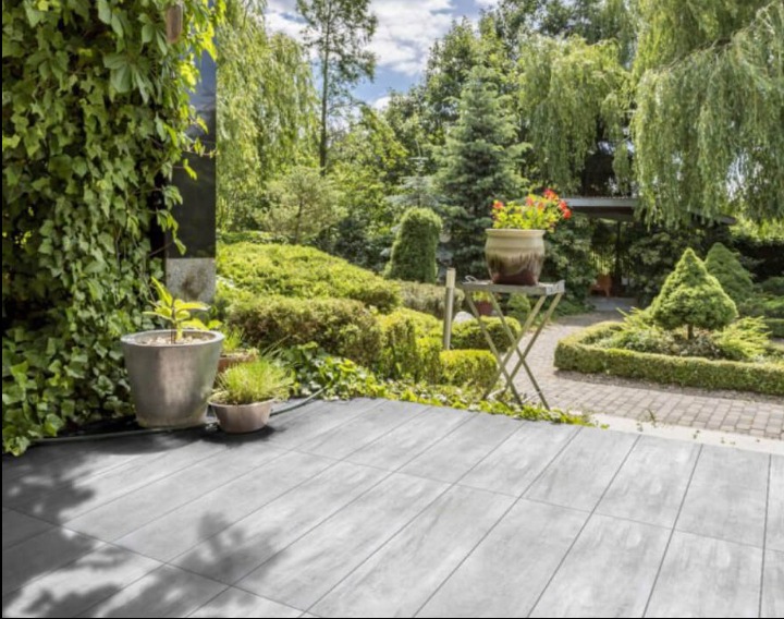 Szare płytki tarasowe ułożone na zewnątrz, z doniczkami z roślinami i widokiem na bujny ogród z drzewami i krzewami.