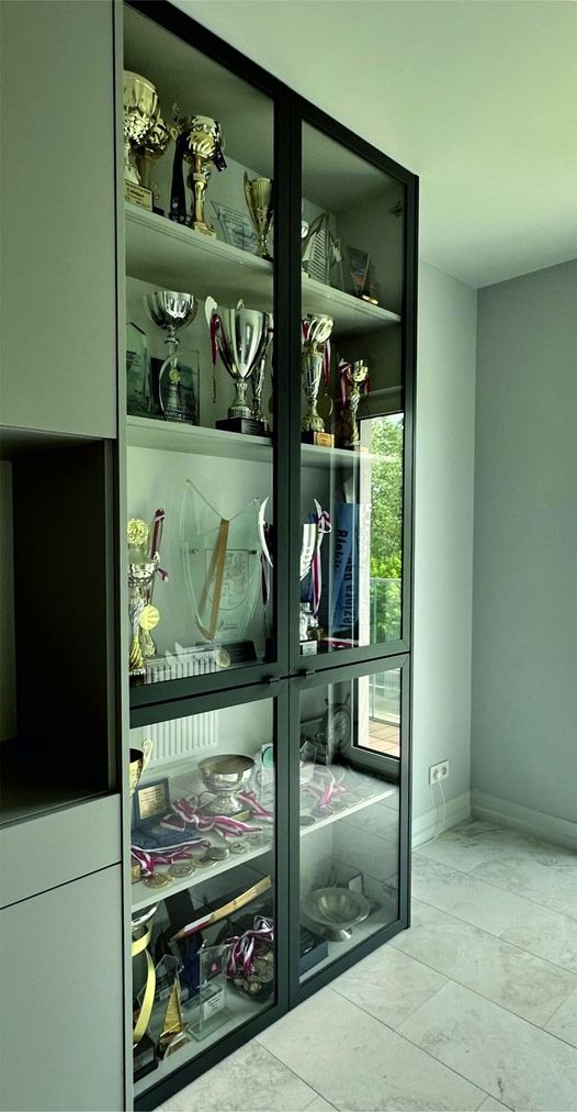 Szklana witryna w zabudowie meblowej, prezentująca kolekcję trofeów sportowych, medali i pucharów na marmurowych półkach.