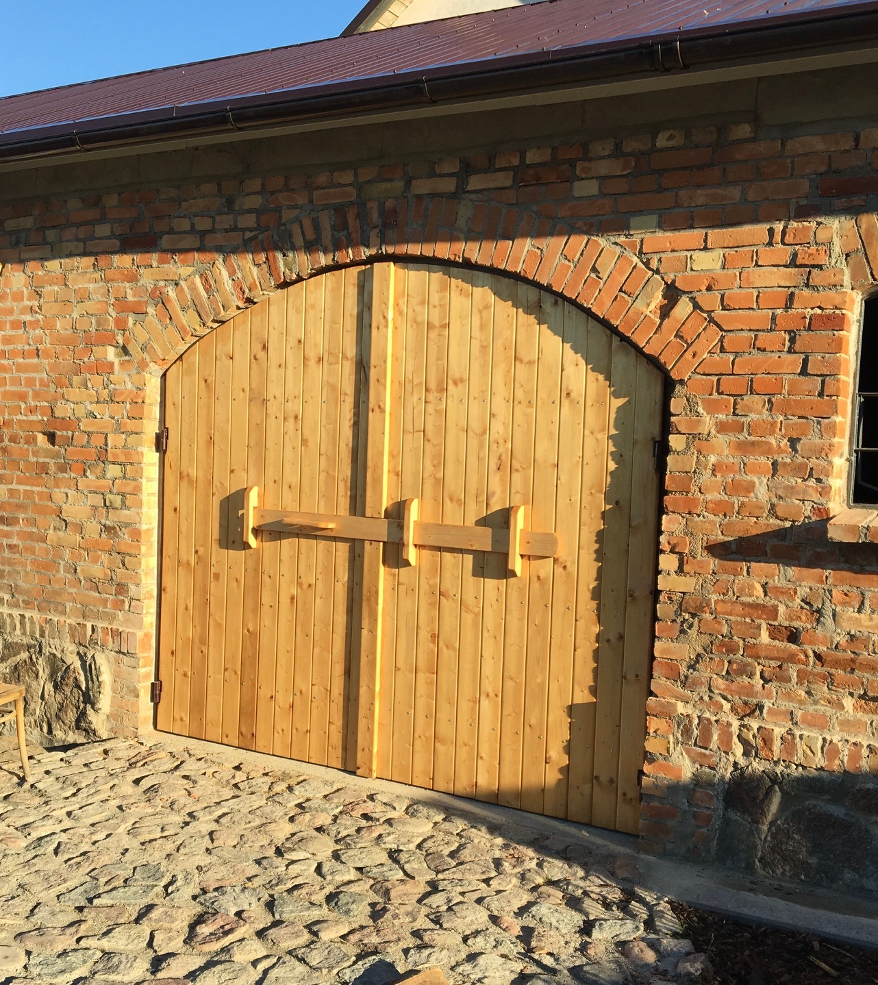 Nowe, dwuskrzydłowe, jasne drewniane drzwi w murze z czerwonej cegły z łukowym nadprożem i prostym drewnianym zamkiem.