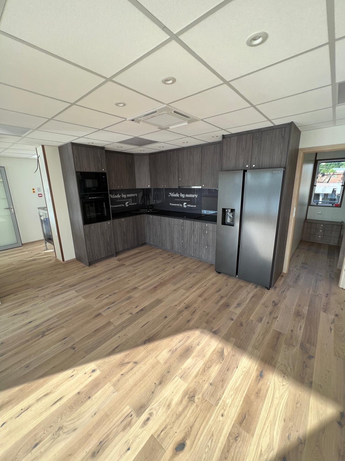 Nowoczesna kuchnia z szarymi drewnopodobnymi frontami, czarnym blatem i szklanym panelem z napisem 'Made by nature', duża lodówka side-by-side, jasna drewniana podłoga, widok z szerokiego kąta.