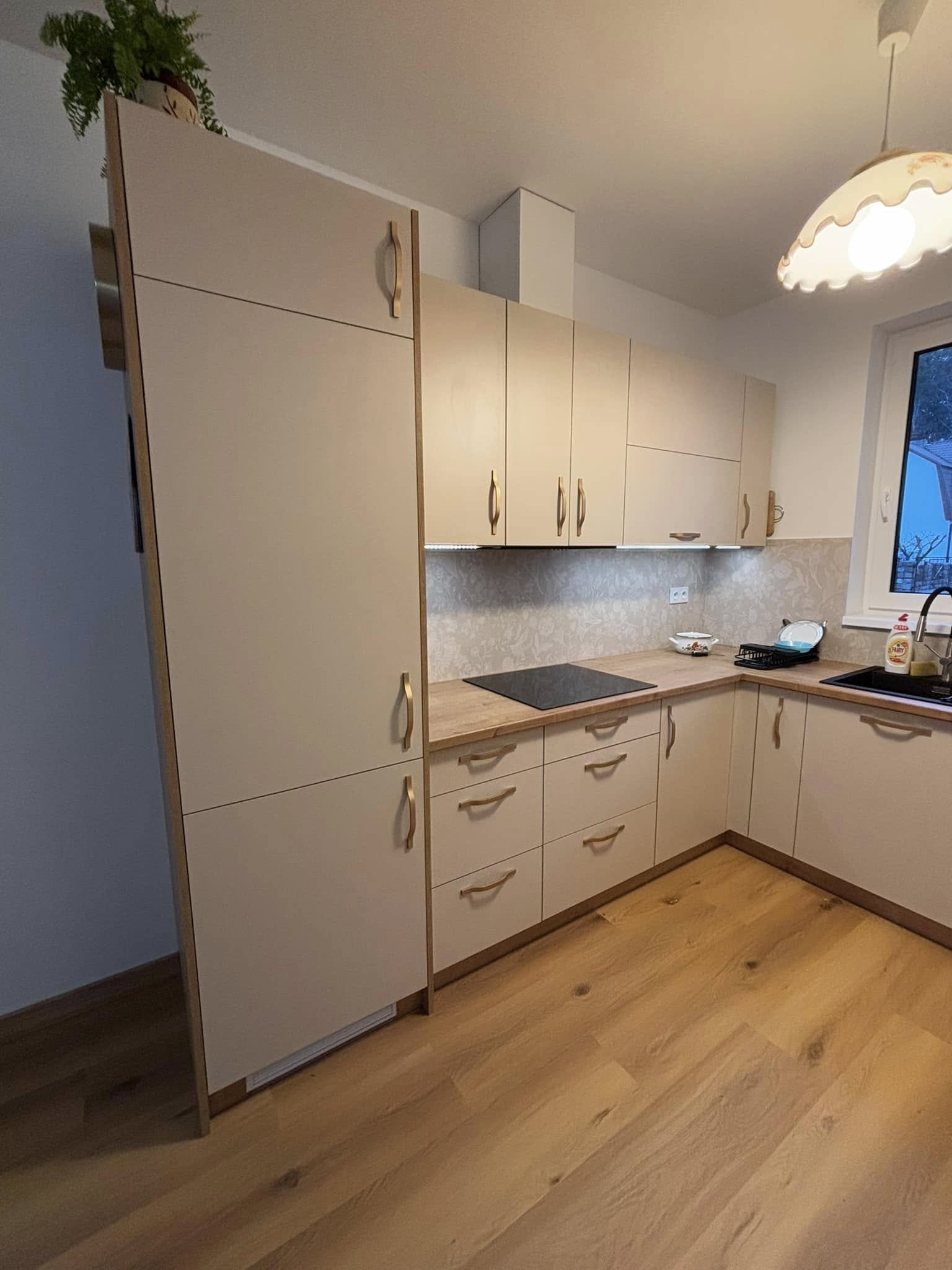 Jasna kuchnia z drewnianymi akcentami, widok na zabudowę z szafkami górnymi i dolnymi, drewniany blat, płyta indukcyjna, zlew i armatura, na górnej szafce doniczka z rośliną.