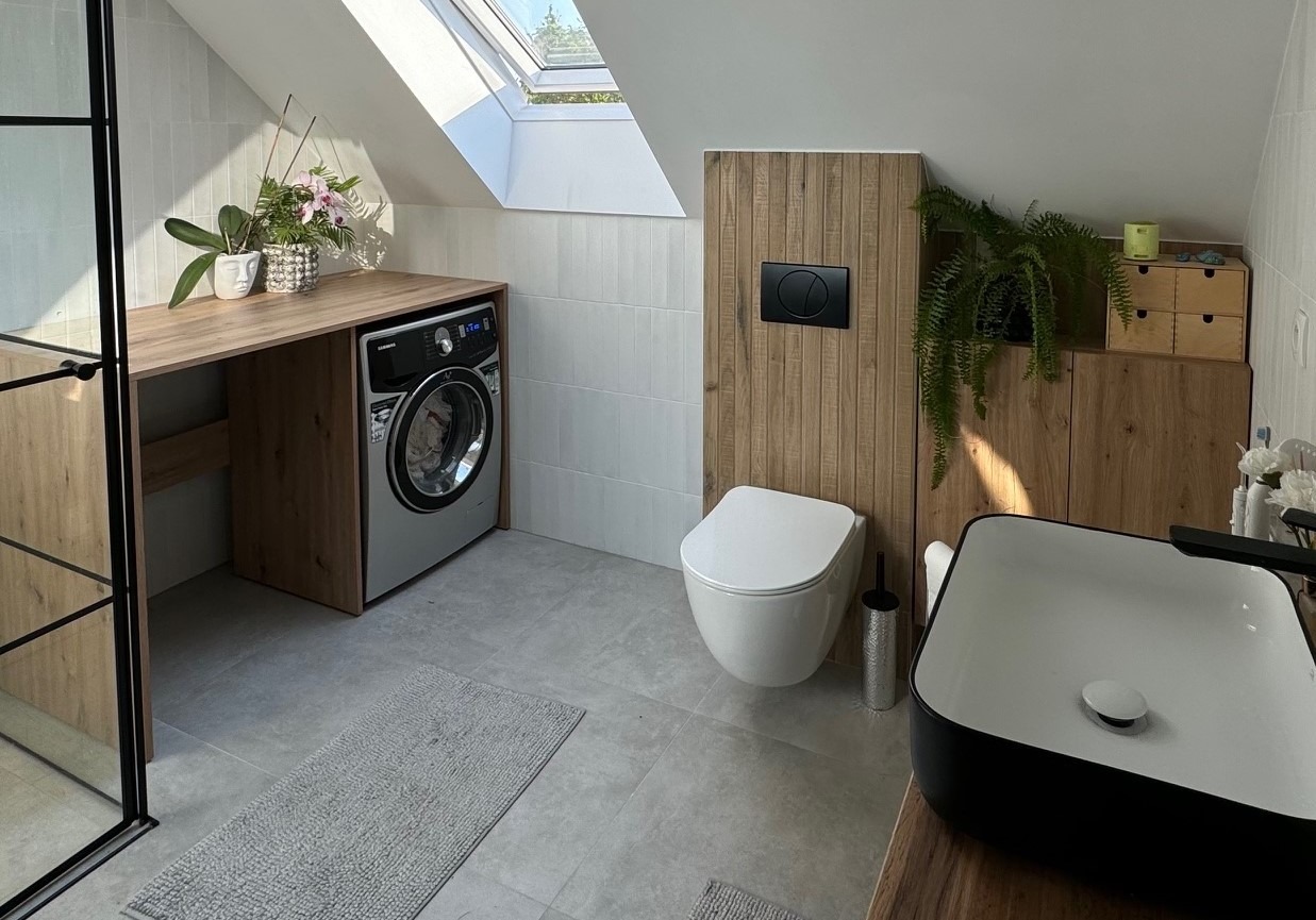 Jasna łazienka na poddaszu ze skośnym sufitem i oknem dachowym. Widoczna zabudowa z drewnianym blatem, pod którym umieszczona jest pralka. Obok toaleta podwieszana i umywalka nablatowa...