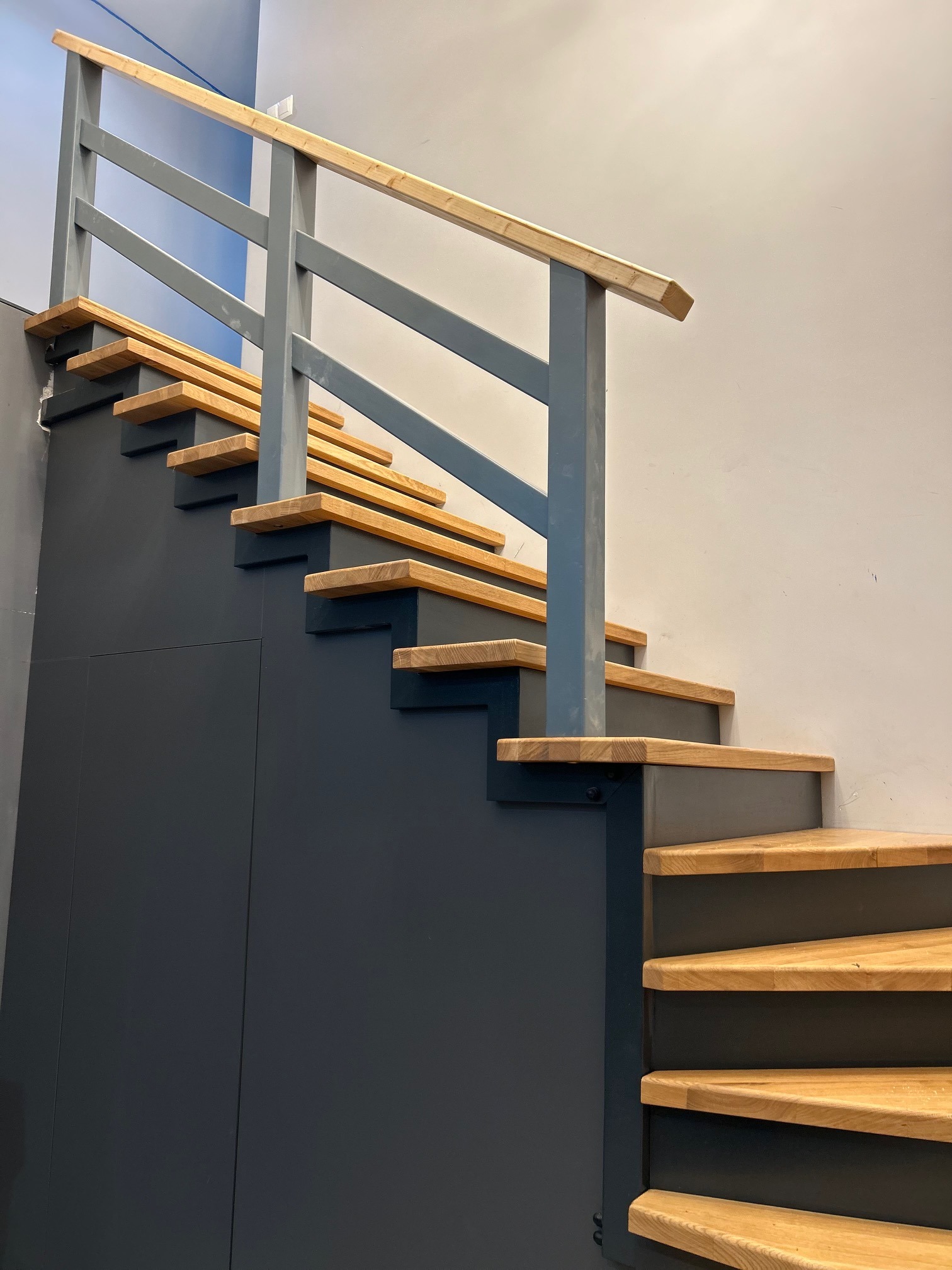 Nowoczesne schody z drewnianymi stopniami i szarą balustradą, częściowo zabudowane szafką w tym samym odcieniu szarości.