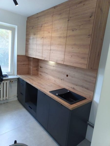 Nowoczesna zabudowa kuchenna z drewnianymi szafkami górnymi i ciemnoszarymi dolnymi, z czarnym zlewem wpuszczanym w blat, oświetlenie podszafkowe, widoczny fragment grzejnika.