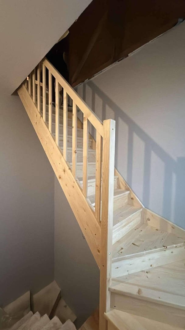 Jasne, sosnowe schody drewniane z prostą balustradą, prowadzące na poddasze z widocznym ociepleniem, przy szarej ścianie.