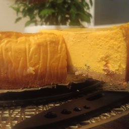 Make with Love for chocolate & white - Kawałek pomarańczowego sernika na kruchym spodzie, podawany na czarnym talerzu z ozdobną podkładką, w tle roślina doniczkowa.