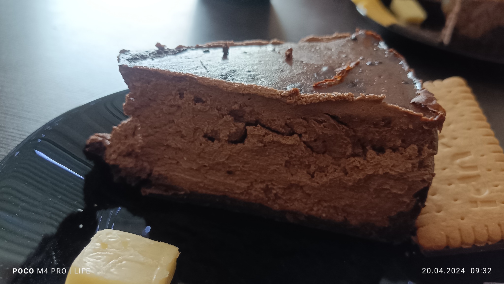 Kawałek czekoladowego sernika na czarnym talerzu, obok kostka masła i herbatnik. Sernik ma błyszczącą polewę.