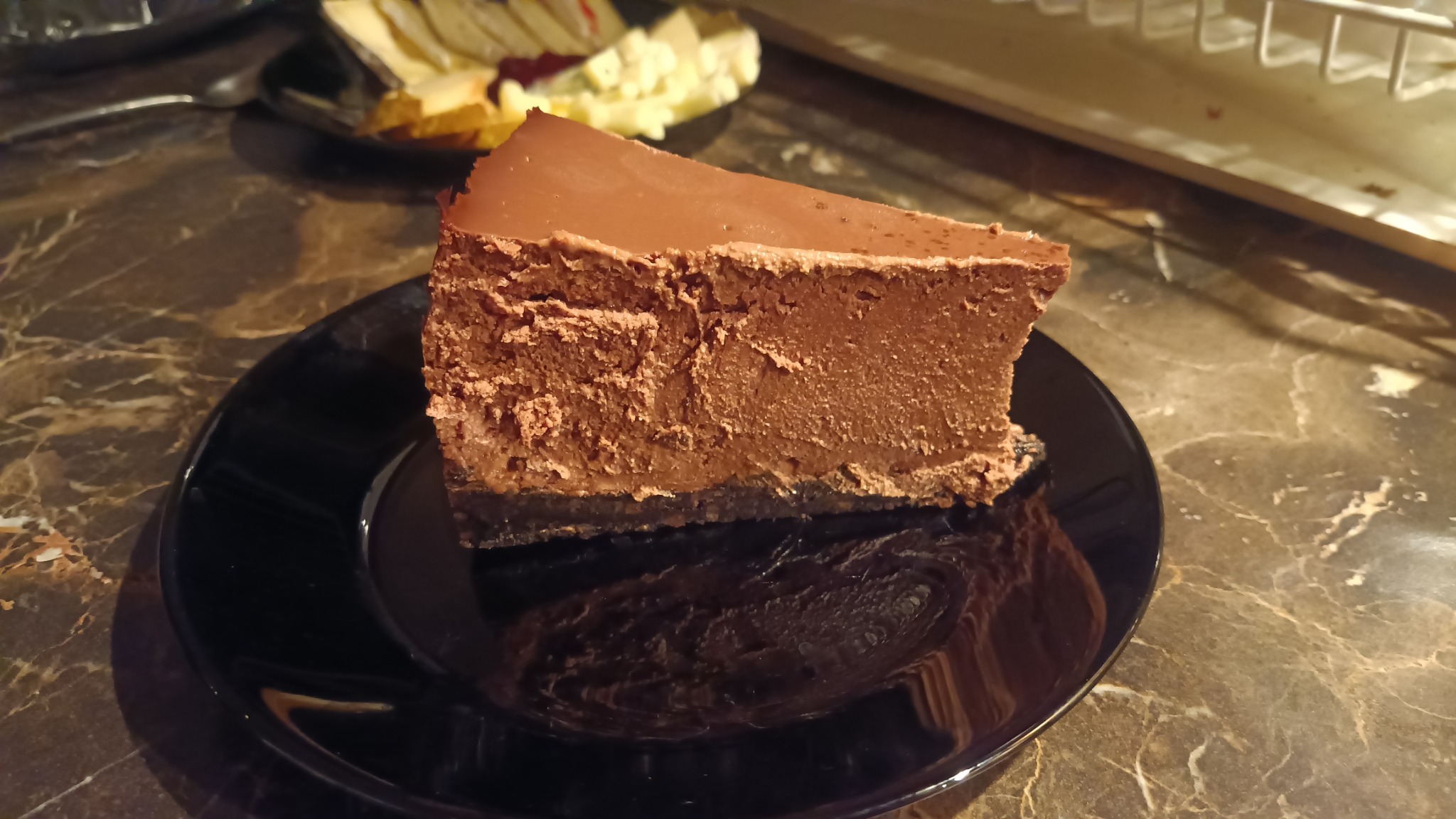 Kawałek czekoladowego sernika z wyraźną warstwą czekoladowej polewy na czarnym talerzu, marmurkowy blat stołu, w tle naczynia do serwowania.