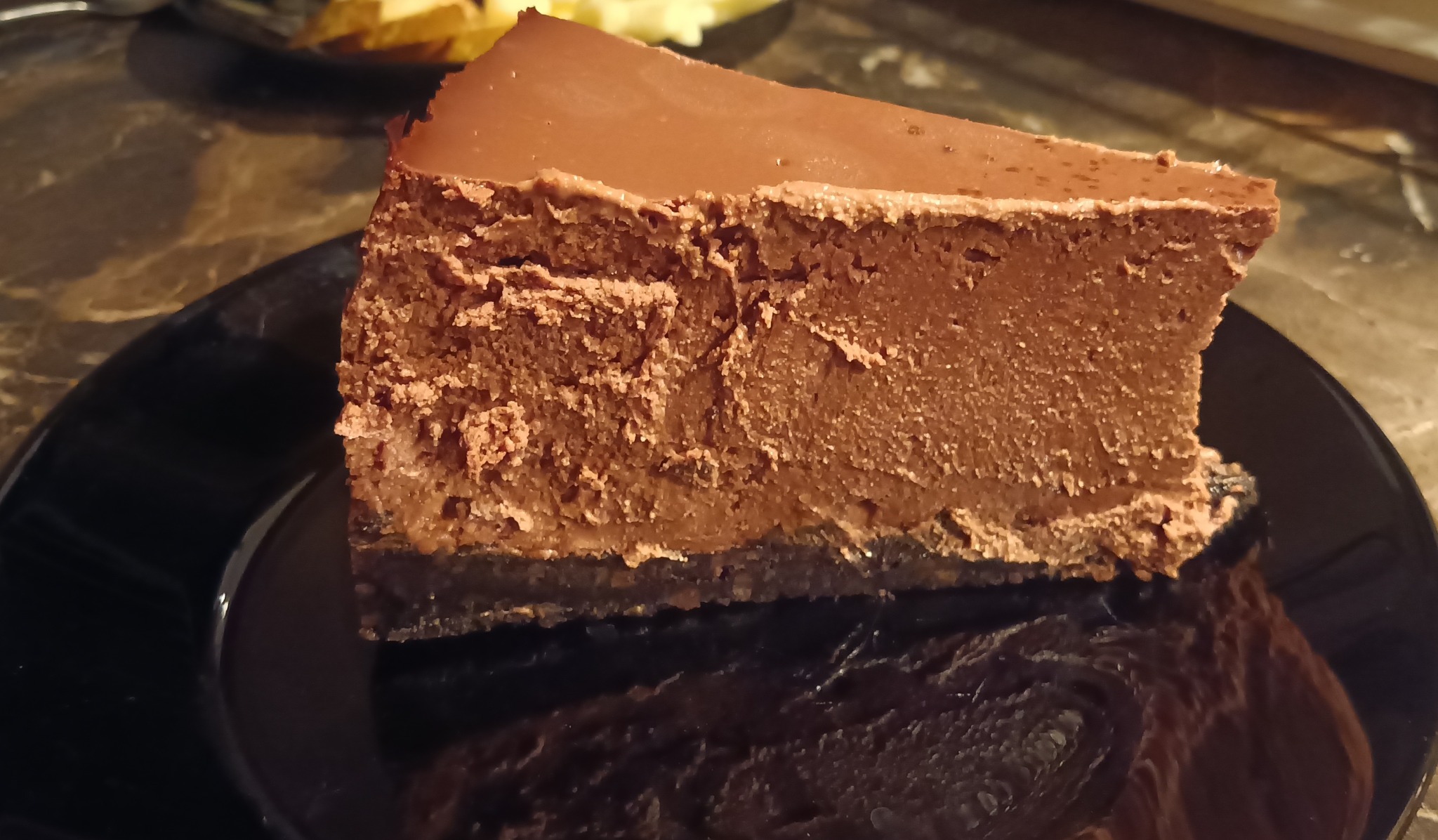 Kawałek czekoladowego ciasta z błyszczącą polewą na czarnym talerzu, widoczne detale tekstury ciasta i odbicie na talerzu.