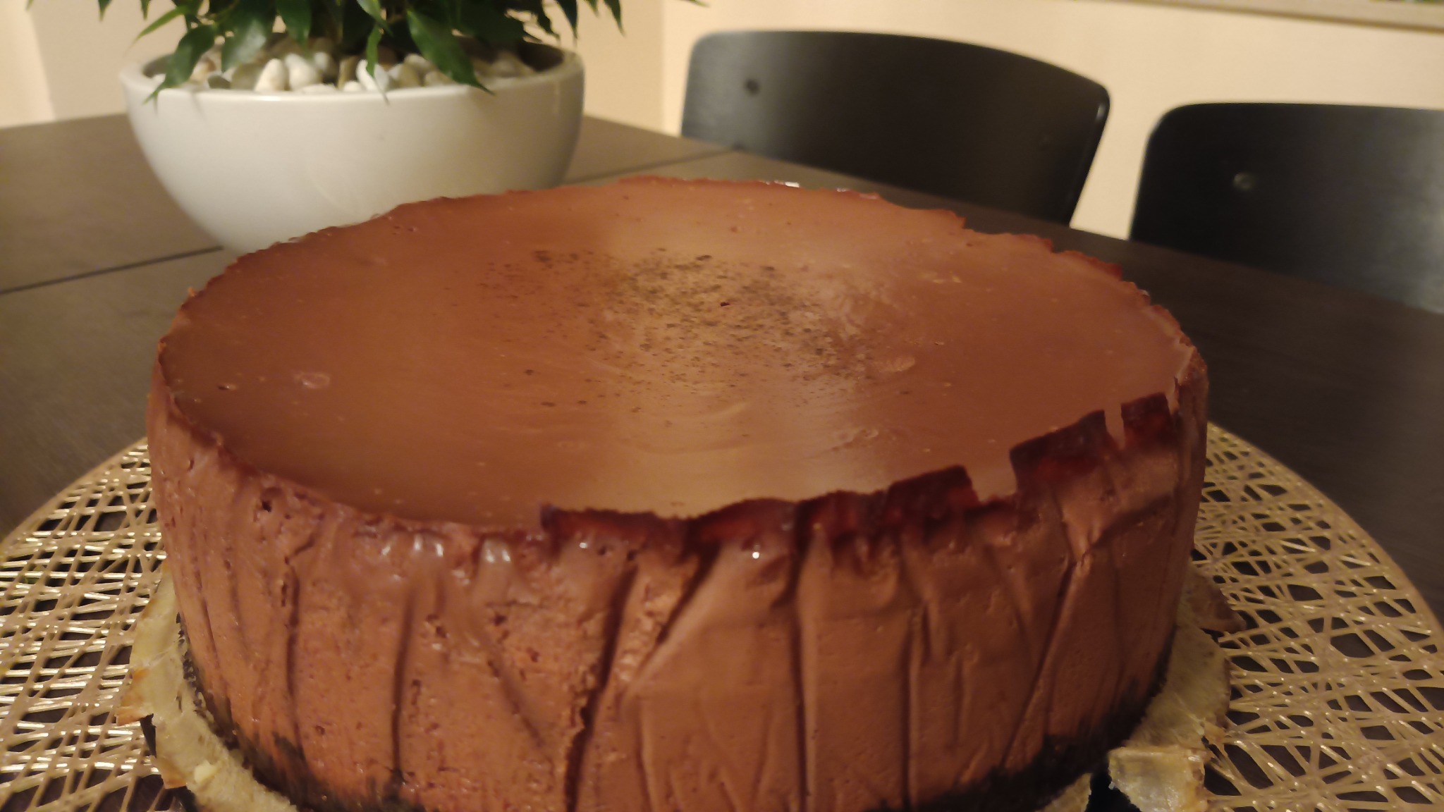 Czekoladowy sernik na spodzie z ciasta, umieszczony na złotej podkładce z plecionki, z rośliną w białej doniczce w tle.