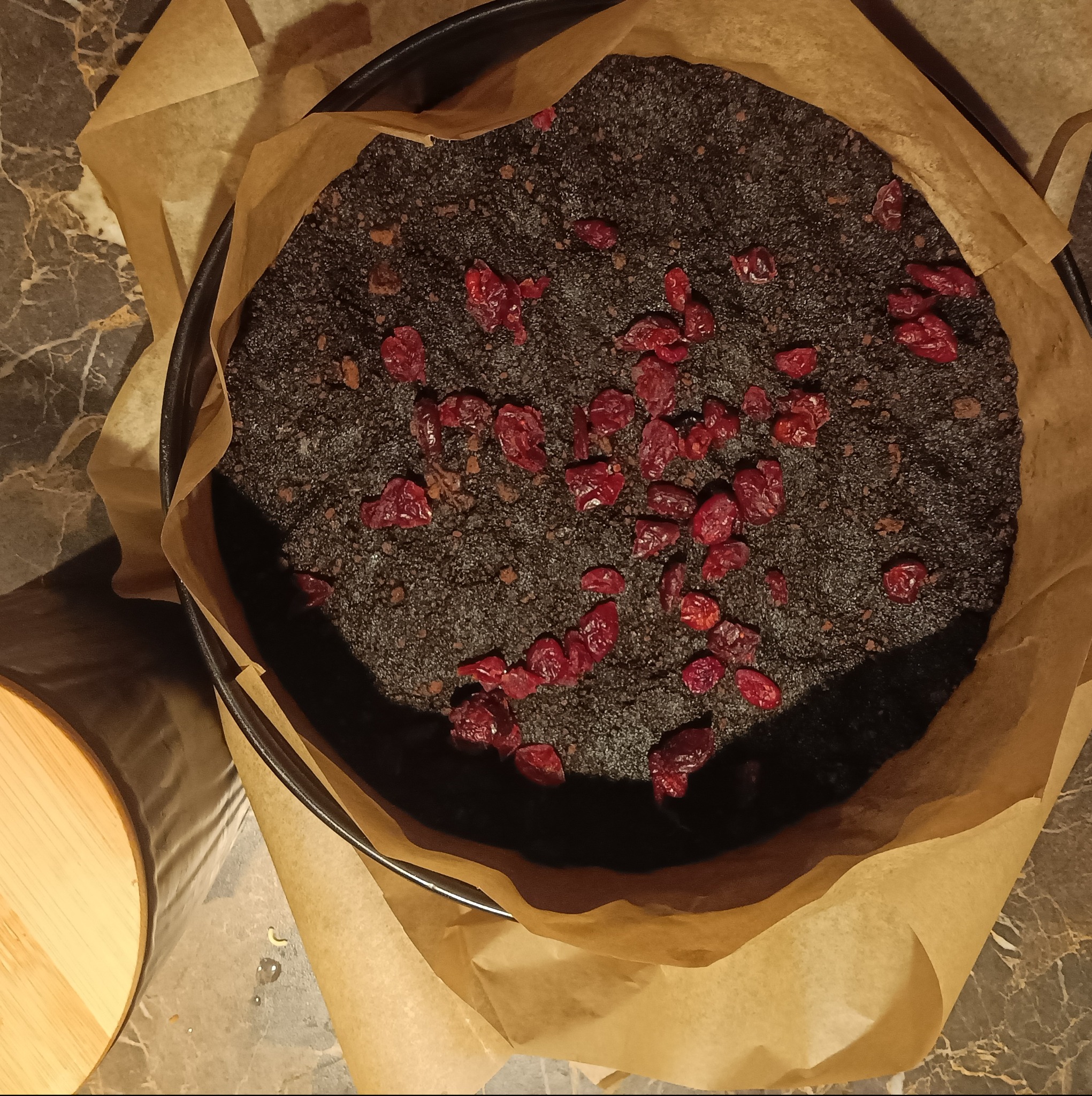 Ciemne ciasto w okrągłej formie wyłożonej papierem do pieczenia, posypane suszoną żurawiną, widoczne na marmurowym blacie.