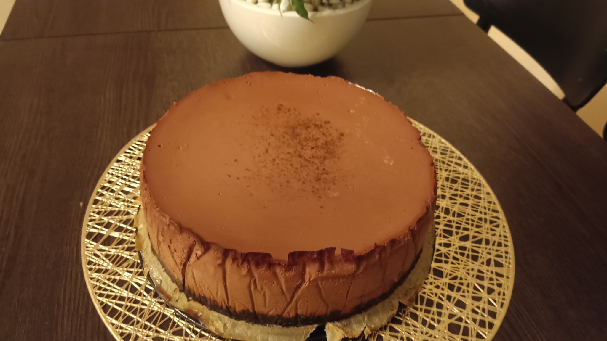 Czekoladowy sernik na złotym podkładzie, delikatnie posypany kakao, ustawiony na ciemnym drewnianym stole z dekoracyjną białą misą w tle.