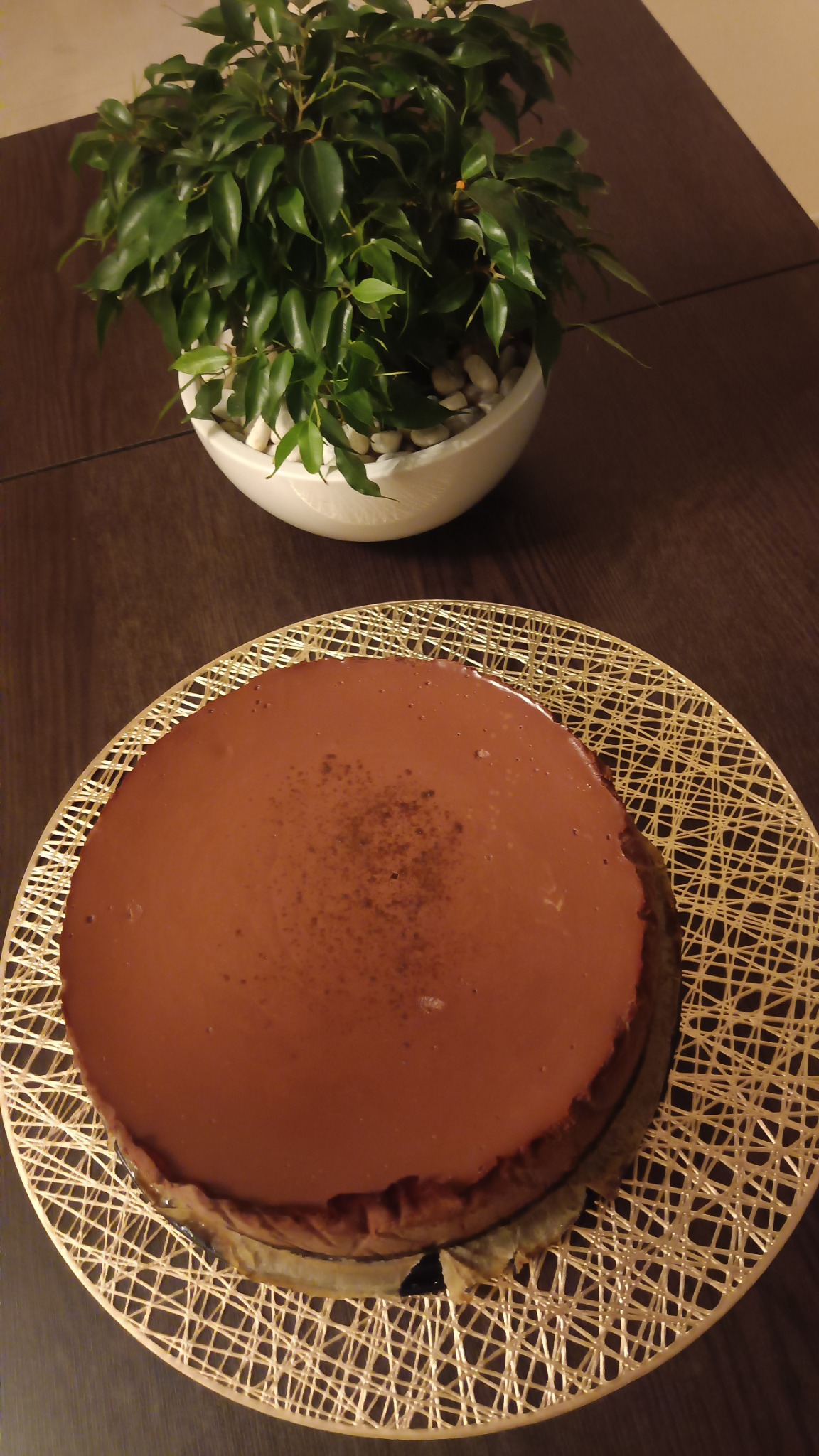 Ciemnobrązowy tort z polewą na ażurowej, okrągłej podkładce, obok doniczka z zieloną rośliną na drewnianym stole.