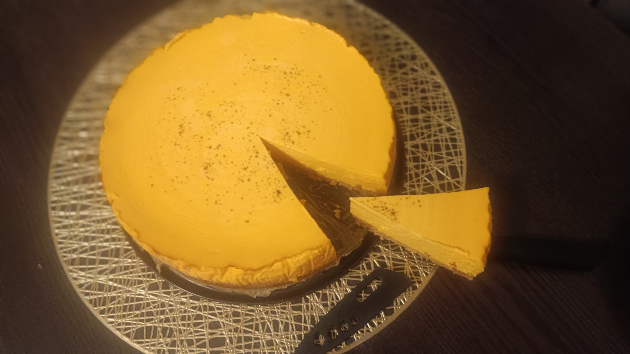 Okazały, okrągły sernik dyniowy z wyciętym kawałkiem, posypany delikatnie przyprawami, na srebrnej, ażurowej podkładce i tle ciemnego blatu.