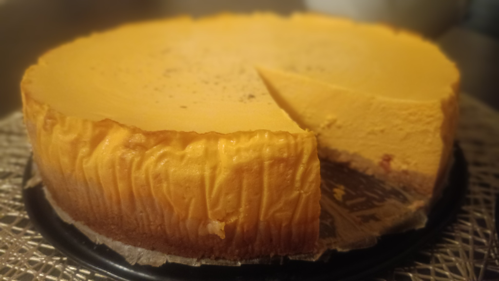 Okazały, pomarańczowy sernik nowojorski z wyciętym kawałkiem, prezentowany na czarnym talerzu, z widoczną strukturą spodu ciasta.