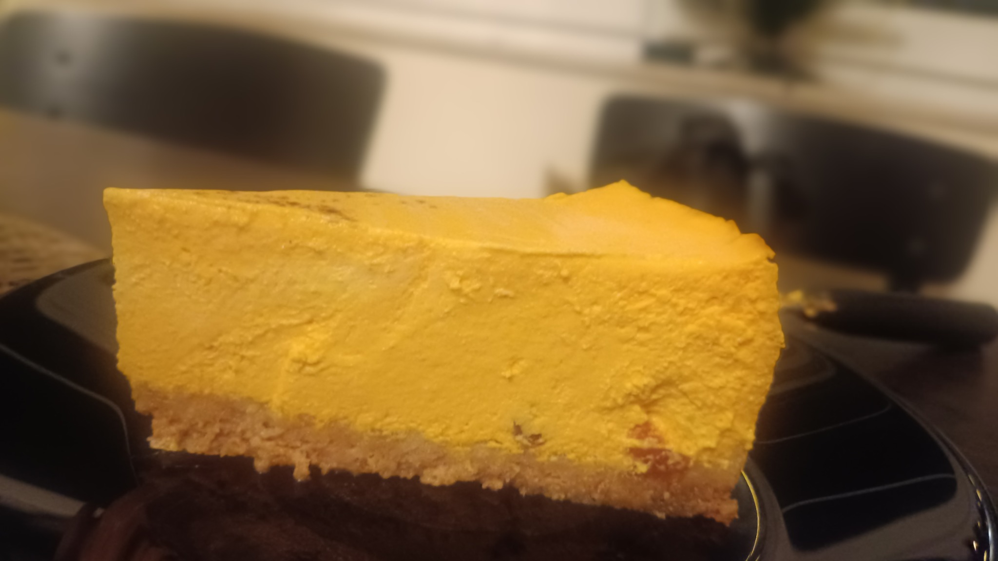 Kawałek sernika o intensywnie żółtym kolorze na czarnym talerzu, z widocznym kruchym spodem i delikatną strukturą wierzchu.